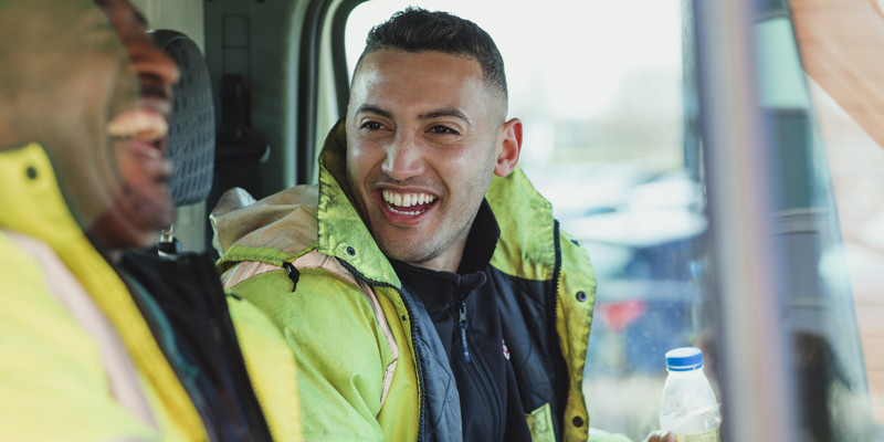 Two Male Labourers Wearing High Vis Jackets Talking And Laughing In Their Work Van Together