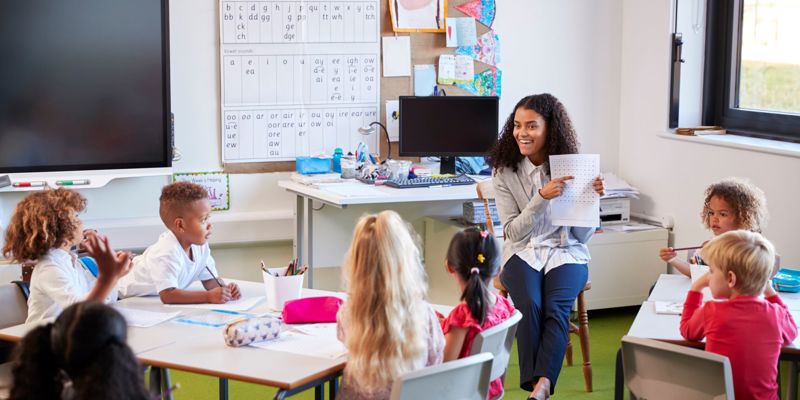 Female primary school teacher sitting on a chair facing school children in a classroom holding up and explaining a worksheet to them